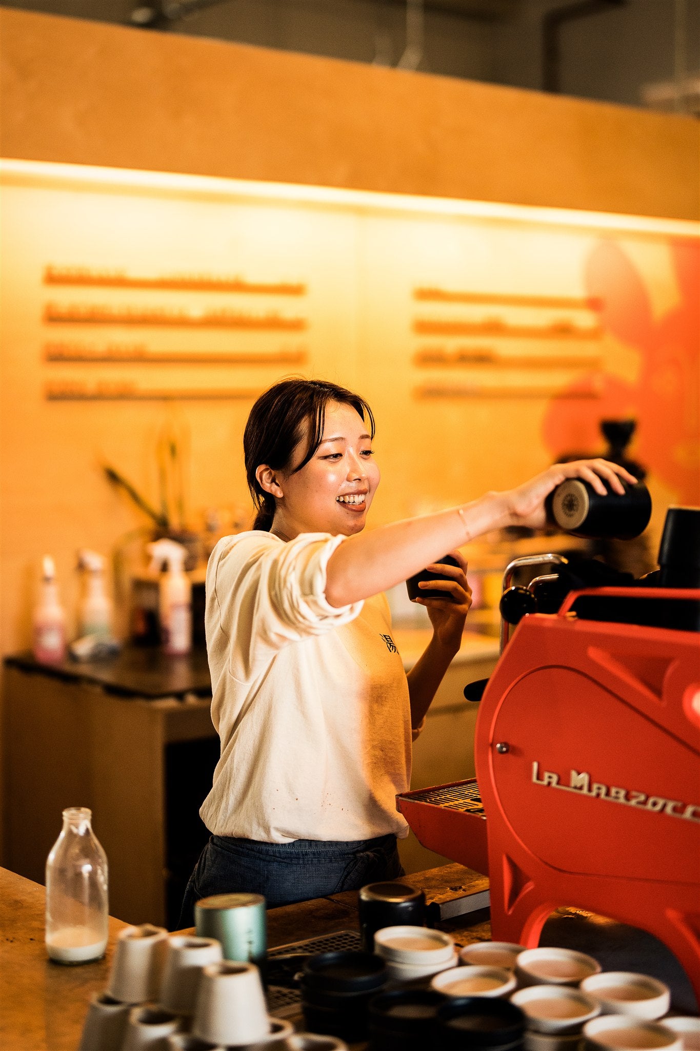 A woman working behind a coffee counter placing a cup down while standing next to a red La Marzocco espresso machine.