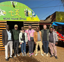 Load image into Gallery viewer, Group of people posing in front of a green sign with text and images on a sunny day.