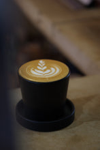 Load image into Gallery viewer, A black ceramic Ritual Coffee branded beaker with saucer on a table, containing a cup of coffee with latte art on top.