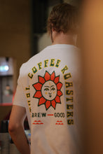 Load image into Gallery viewer, A person wearing a Ritual Coffee branded white crew neck t-shirt with a green and red sun design and the text 'COFFEE ROASTERS BREW GOOD'.