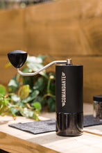 Load image into Gallery viewer, A black Comandante C40 Nitro Blade Grinder with a silver crank, placed on a table with a plant in the background.