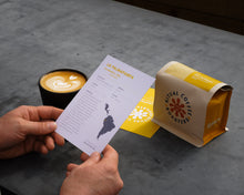 Load image into Gallery viewer, Person holding an information card on La Palmapampa coffee. In the background there is a cup of coffee and a bag of the coffee with the Ritual Coffee logo on it